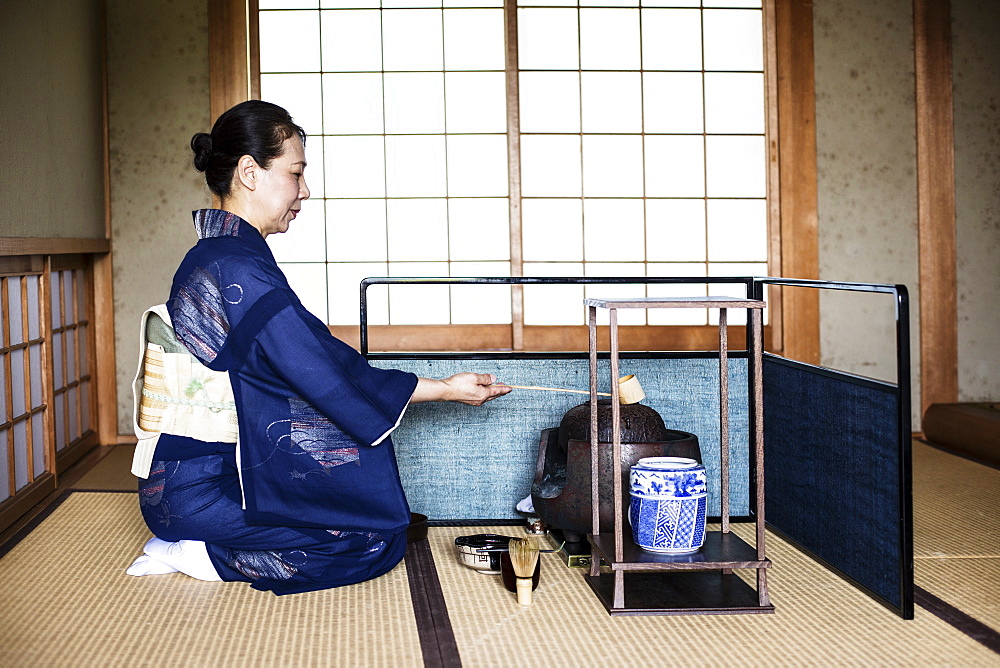 Japanese woman wearing traditional bright blue kimono with cream coloured obi kneeling on floor, using a  Hishaku, a bamboo ladle, during a tea ceremony, Kyushu, Japan