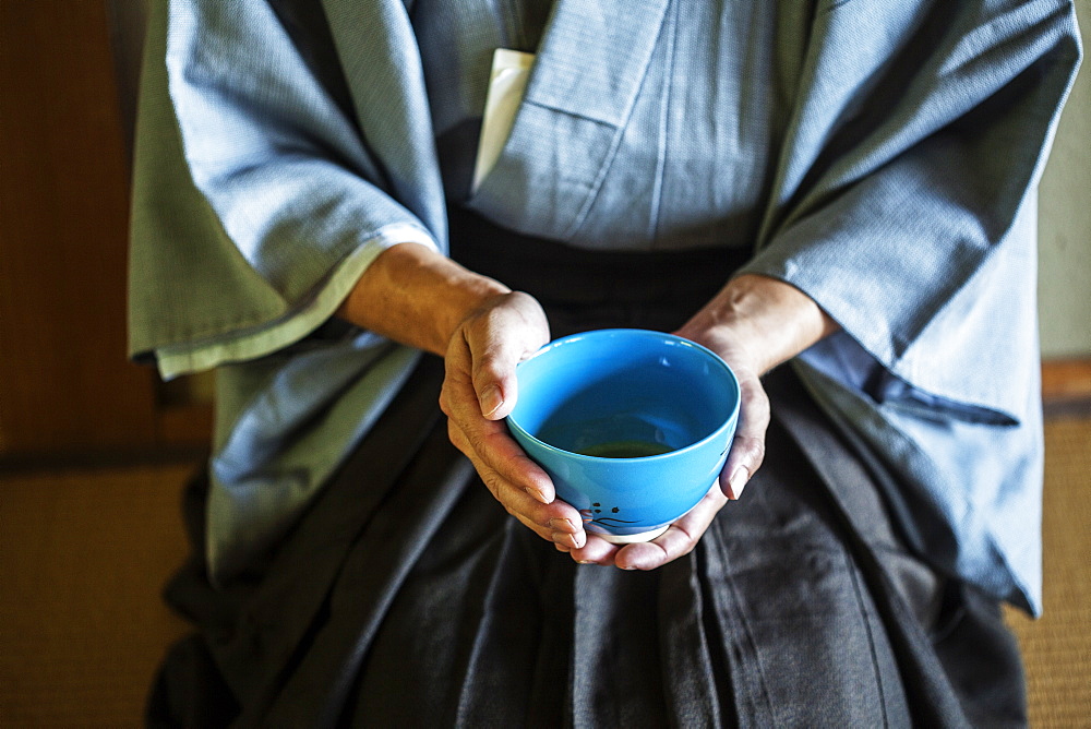 High angle close up of Japanese man wearing traditional kimono kneeling on floor holding blue tea bowl during tea ceremony, Kyushu, Japan