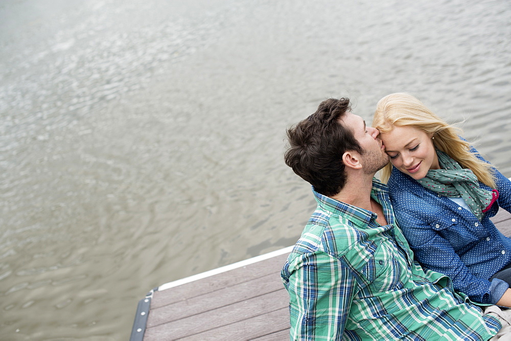 A man and woman seated on a jetty by a lake.