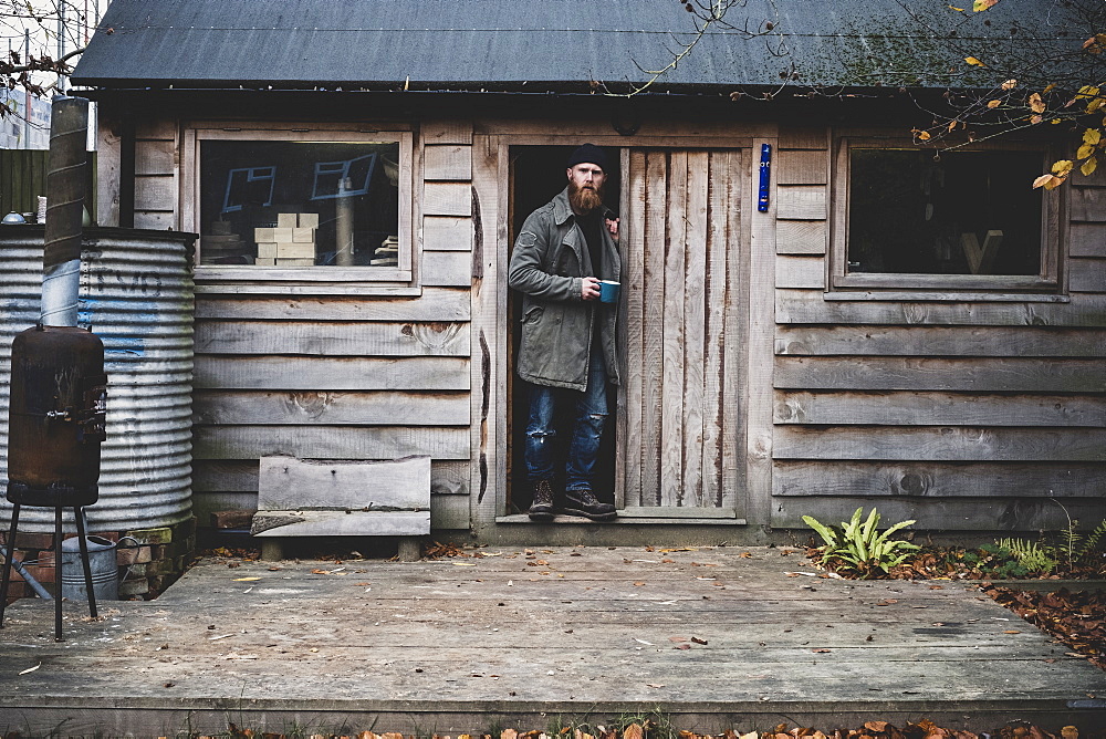 Bearded man standing in doorway of wooden workshop, holding blue mug, looking at camera, Berkshire, England