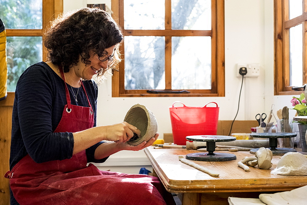 Woman wearing red apron sitting in her ceramics workshop, working on small clay bowl, England