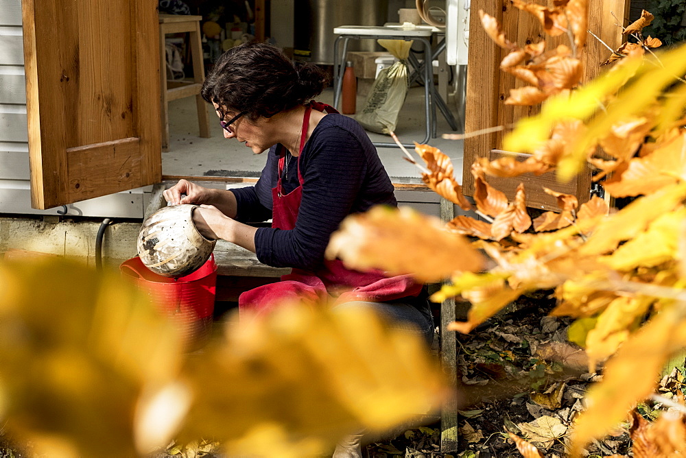 Woman wearing red apron sitting outside her ceramics workshop, holding vase, England