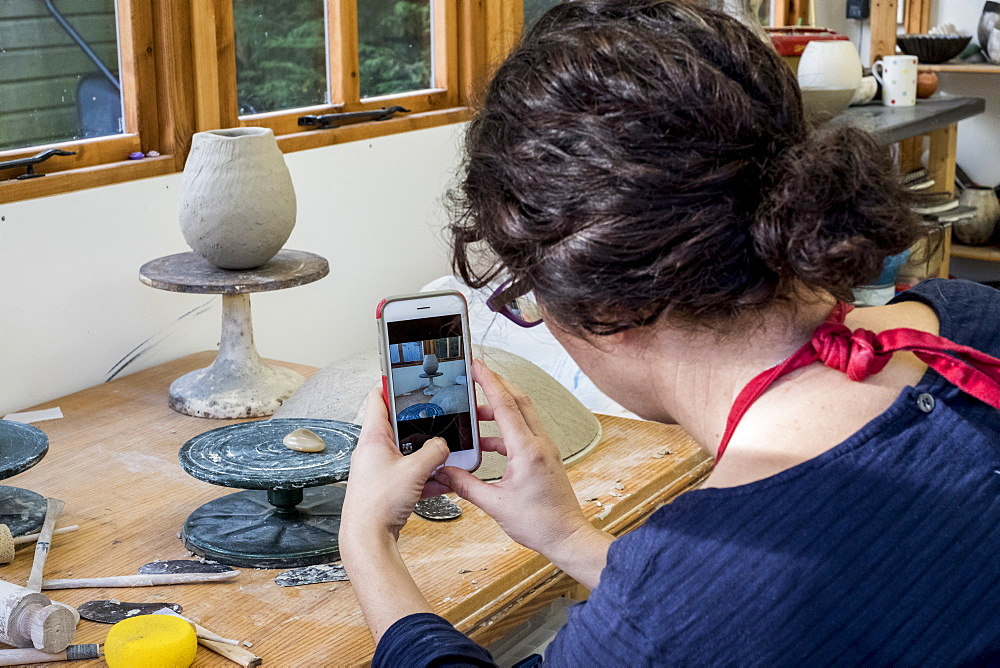 Over the shoulder view of woman sitting in her ceramics workshop, checking her mobile phone, England