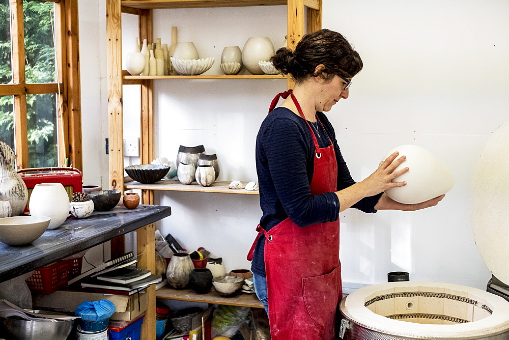 Woman wearing red apron standing in her workshop next to kiln, holding ceramic vase, England