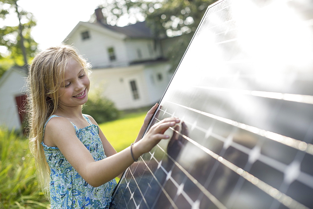 A young girl beside a large solar panel in a farmhouse garden. 