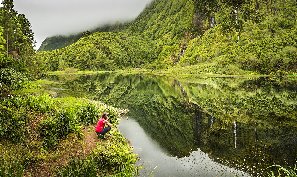 Hiker crouching at still lake in remote mountains, Faja Grande, Flores, Portugal