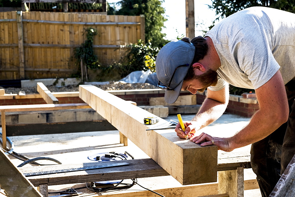 Man wearing baseball cap standing on building site, measuring and marking wooden beam, Oxfordshire, England