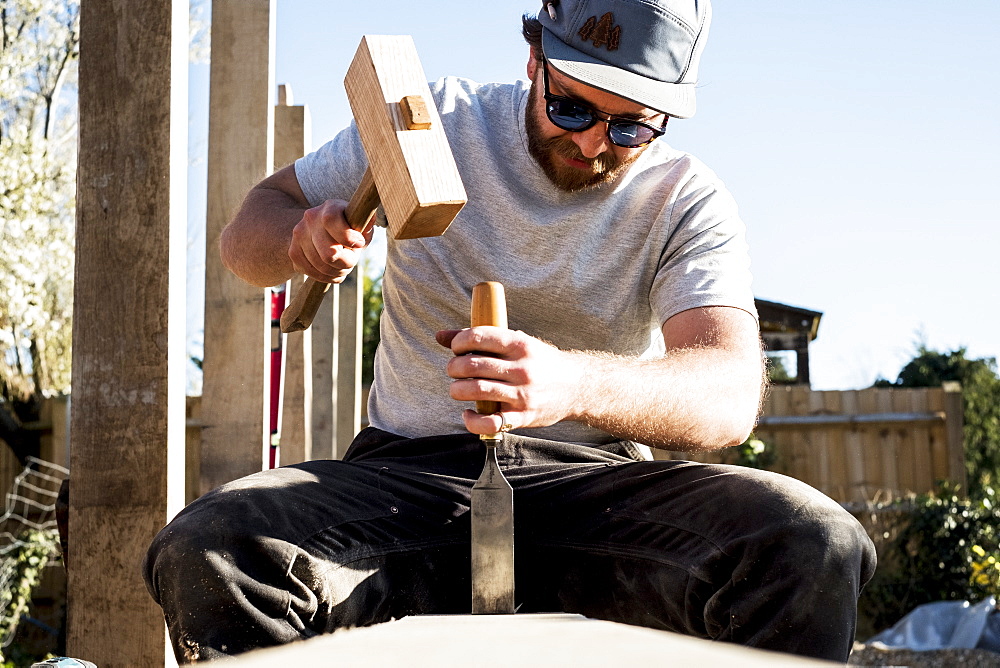 Man wearing baseball cap and sunglasses on building site, using mallet and chisel, working on wooden beam, Oxfordshire, England