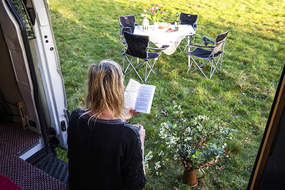 High angle rear view of woman sitting in door of camper van parked on a meadow, reading a book, Oxfordshire, England