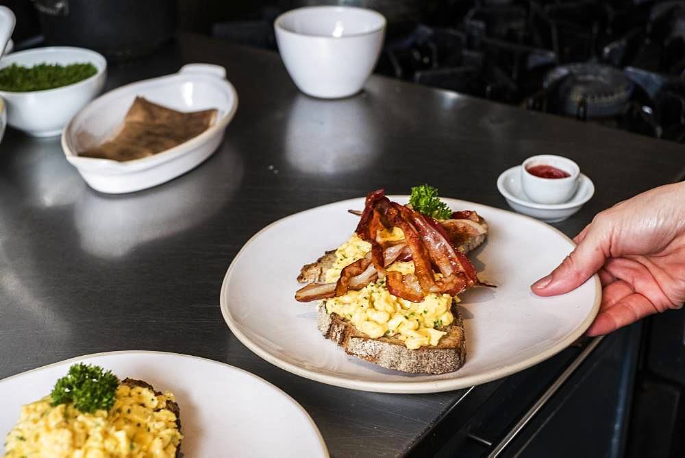 High angle close up of person holding plate with scrambled eggs and crispy bacon on a slice of bread