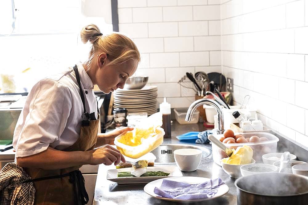 Female chef wearing brown apron standing at a kitchen counter, garnishing plate of asparagus