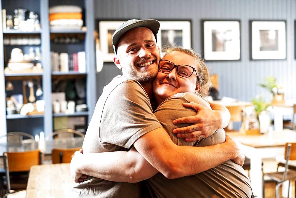 Man wearing baseball cap and woman wearing glasses hugging, smiling at camera