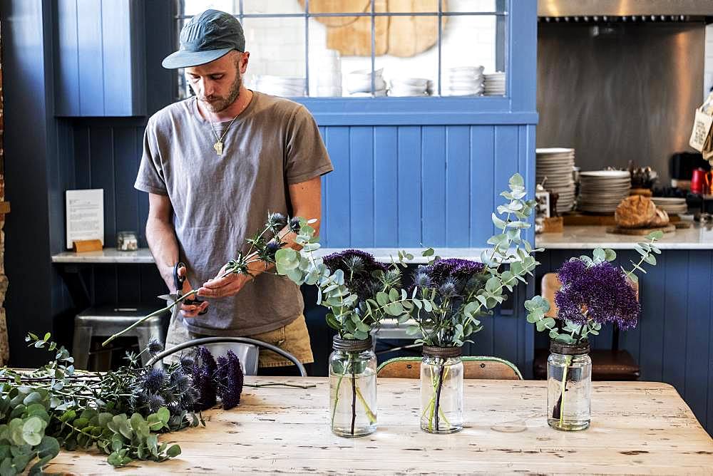 Man wearing baseball cap standing at a table, cutting thistles for flower arrangements in large glass jars