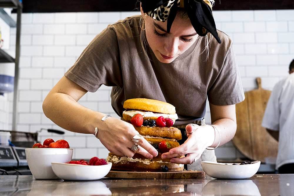 A cook working in a commercial kitchen assembling a layered sponge cake with fresh fruit