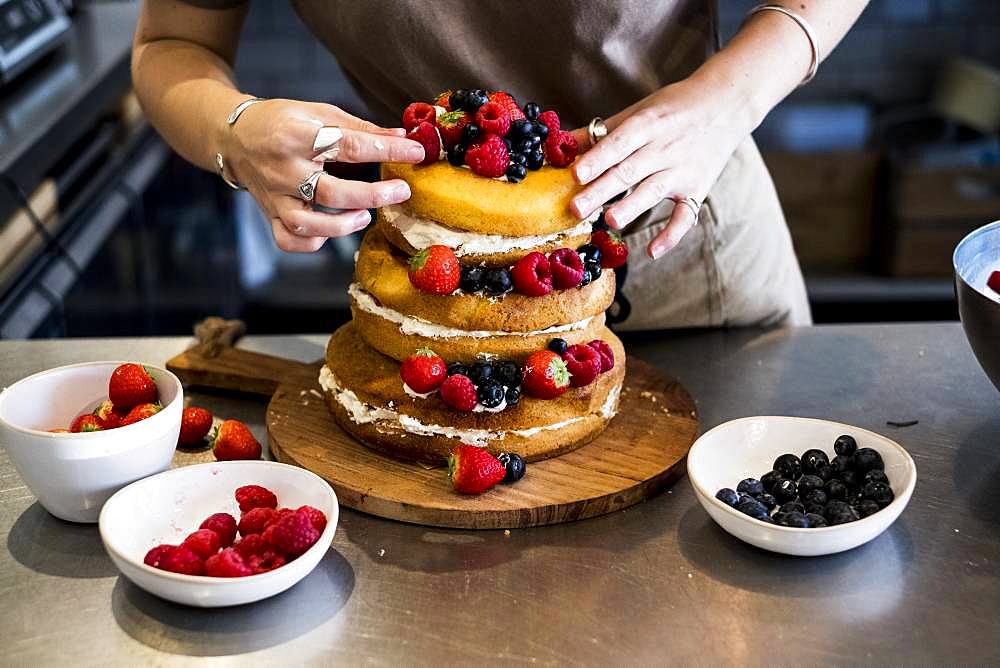 a cook working in a commercial kitchen arranging fresh fruit over a layered cake with fresh cream