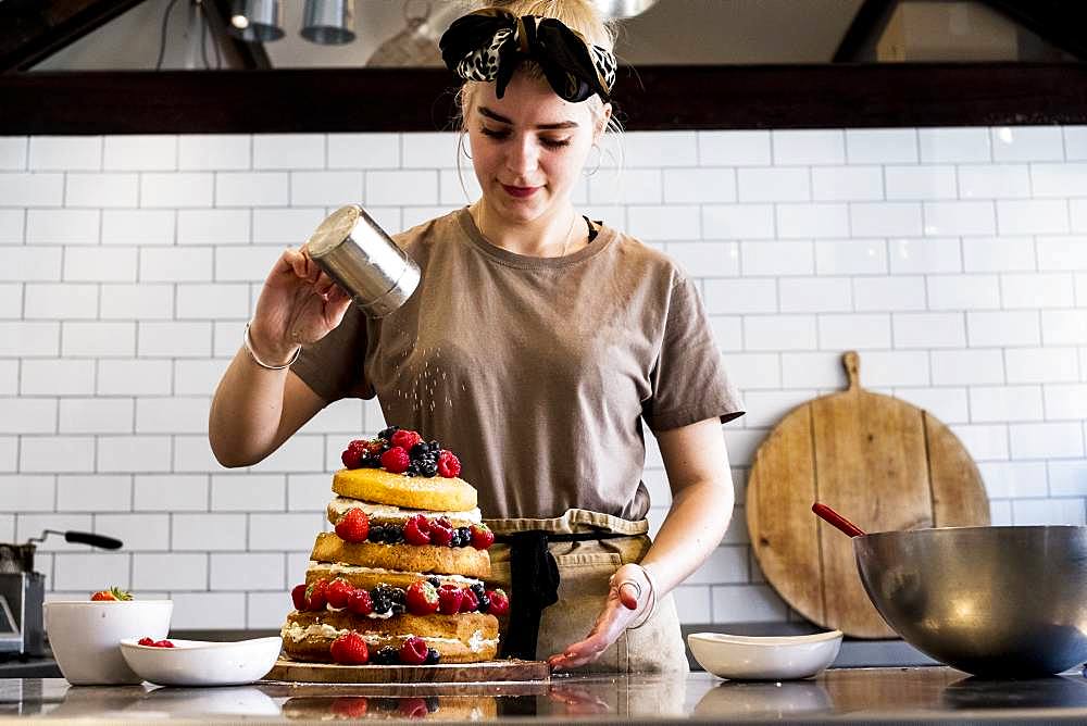 A cook working in a commercial kitchen sprinkling icing sugar over a layered cake with fresh fruit