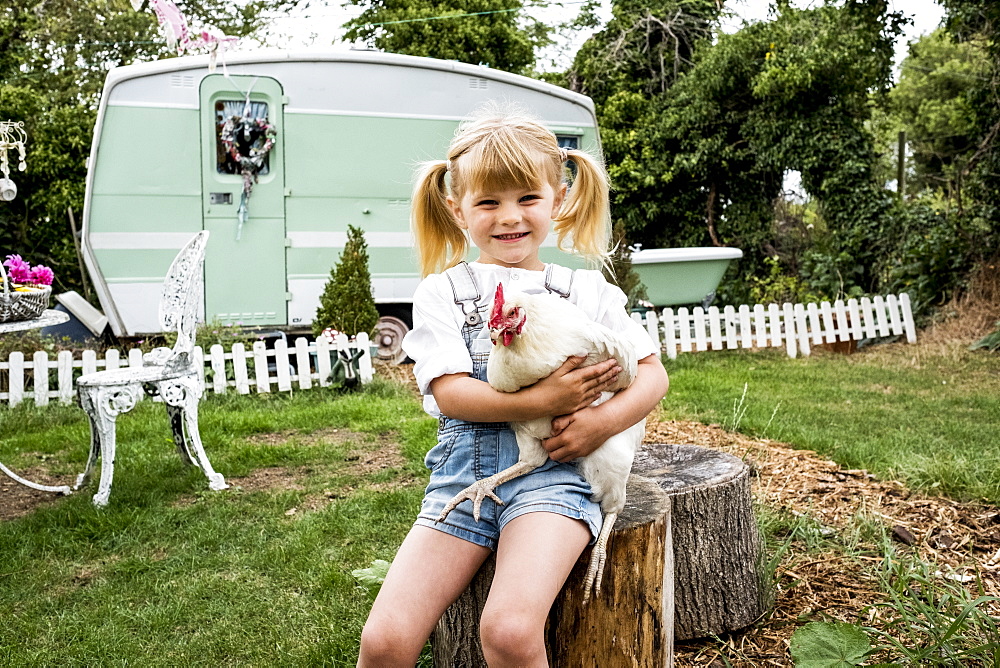 Blond girl sitting in garden holding white chicken, white and green retro caravan in background