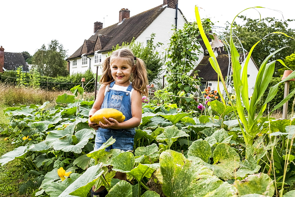 Girl standing in a vegetable patch in a garden, holding yellow gourd, smiling at camera