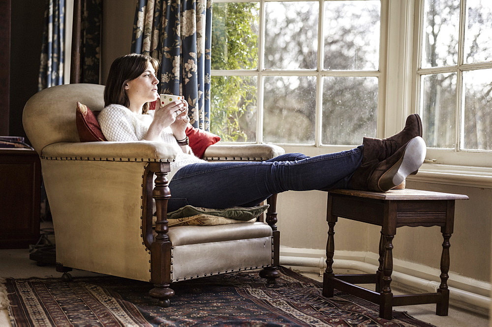 A young woman relaxing at home, with her feet up, having a cup of tea, Ringwood, Hampshire, England