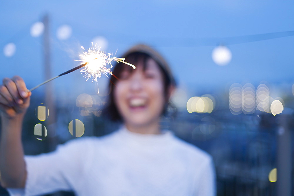 Smiling young Japanese woman holding sparkler on a rooftop in an urban setting, Fukuoka, Kyushu, Japan