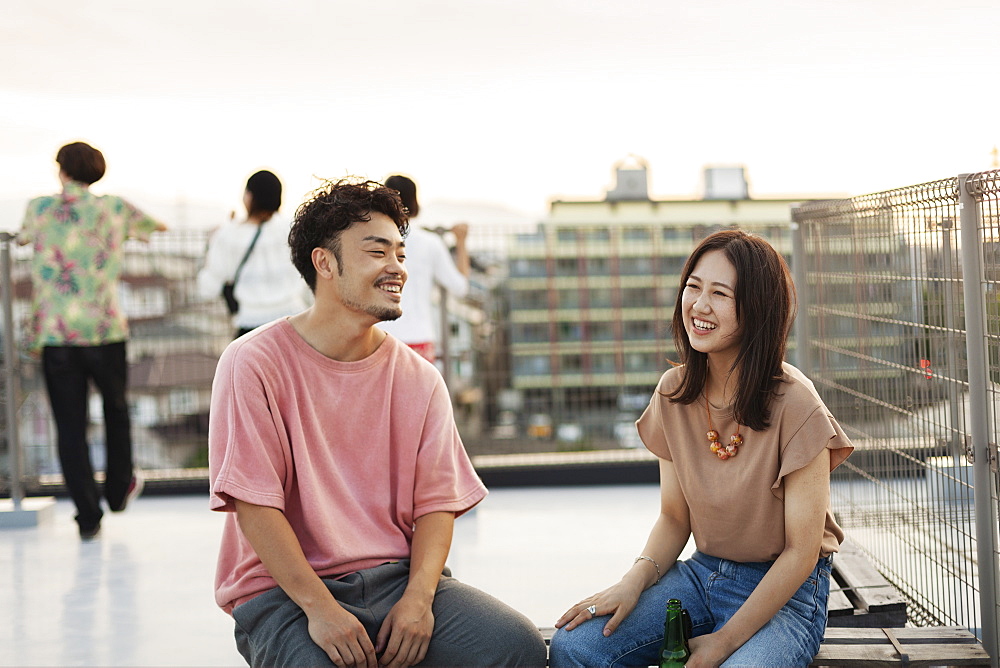 Smiling young Japanese man and woman sitting on a rooftop in an urban setting, Fukuoka, Kyushu, Japan