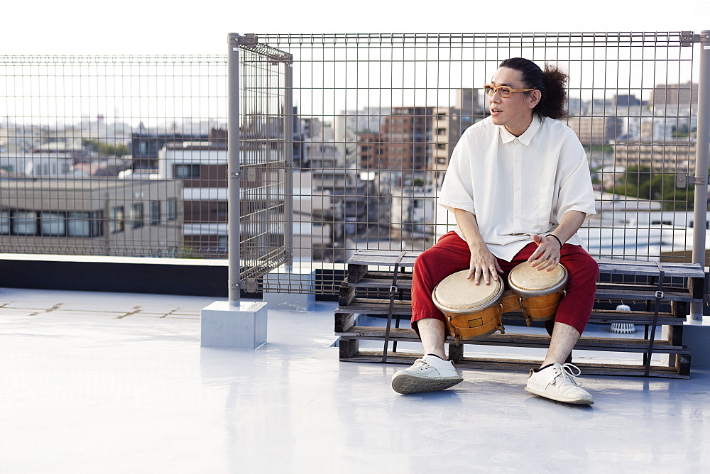 Japanese man sitting on a rooftop in an urban setting, playing drums, Fukuoka, Kyushu, Japan