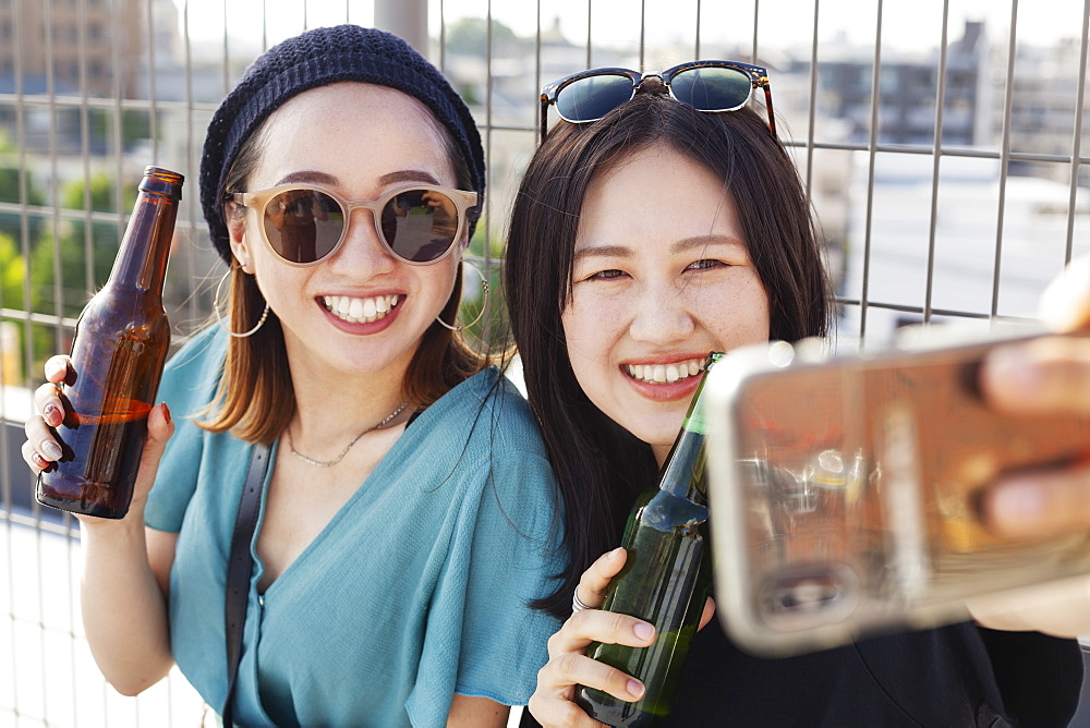 Two young Japanese women sitting on a rooftop in an urban setting, taking selfie with mobile phone, Fukuoka, Kyushu, Japan