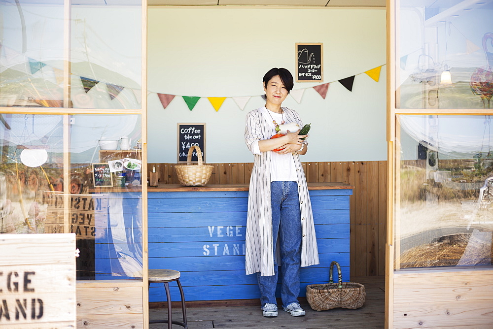 Japanese woman standing in a farm shop, holding vegetables, smiling at camera, Kyushu, Japan