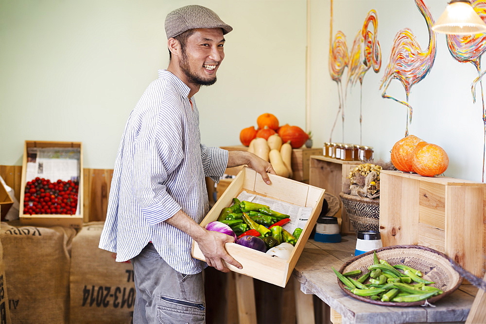 Smiling Japanese man wearing cap standing in farm shop, holding crate with fresh vegetables, Kyushu, Japan