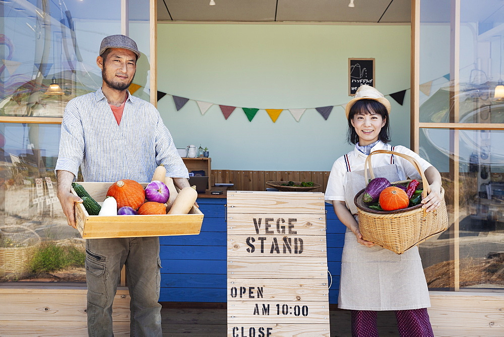 Japanese man and woman standing outside a farm shop, holding crate and basket with fresh vegetables, looking at camera, Kyushu, Japan