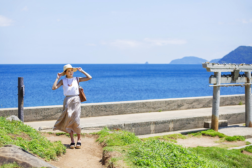 Japanese woman wearing hat walking along path by the ocean, Kyushu, Japan