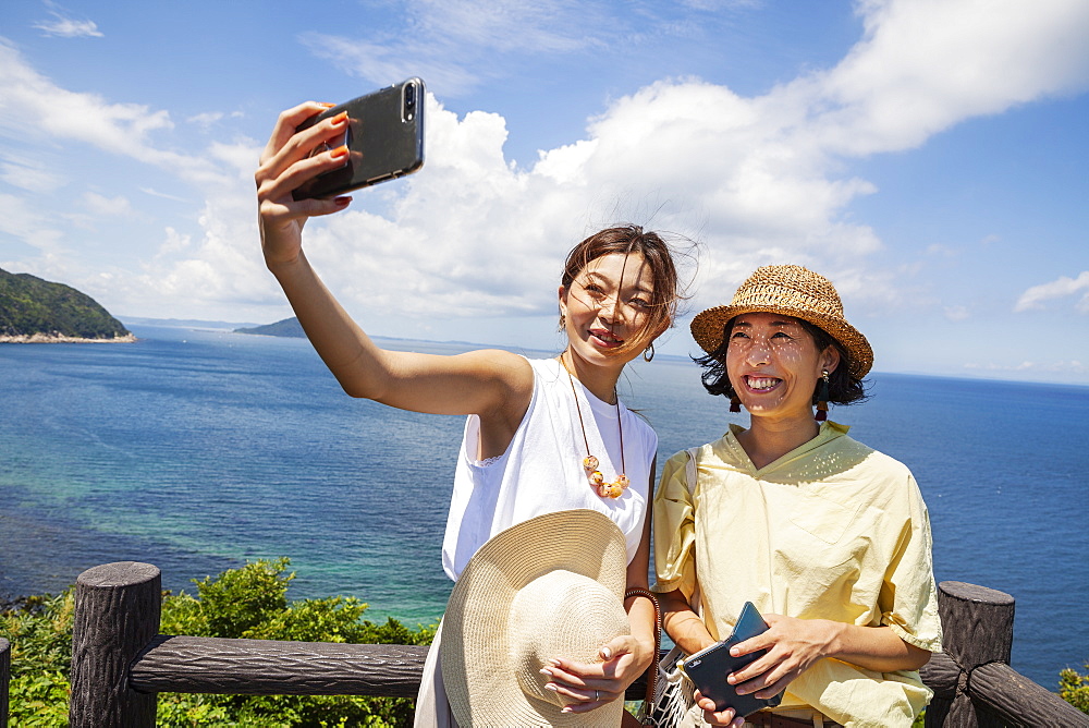 Two Japanese women wearing hats standing on a cliff, taking selfie with mobile phone, ocean in the background, Kyushu, Japan