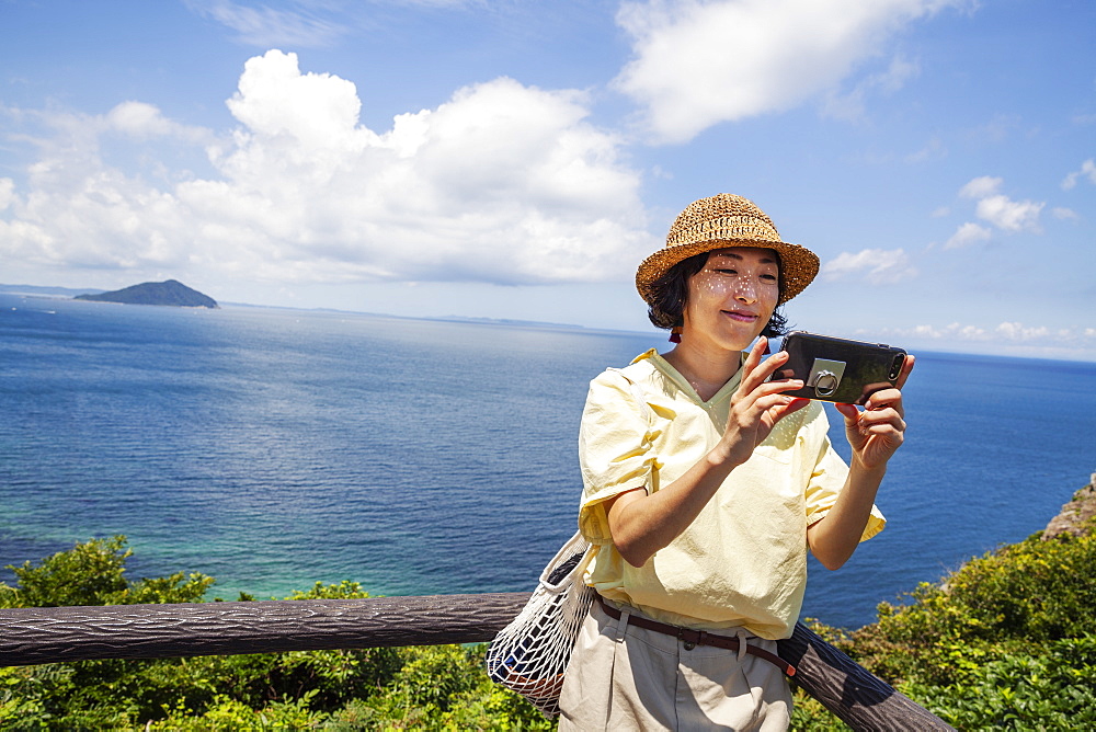 Japanese woman wearing hat standing on a cliff, taking selfie with mobile phone, ocean in the background, Kyushu, Japan