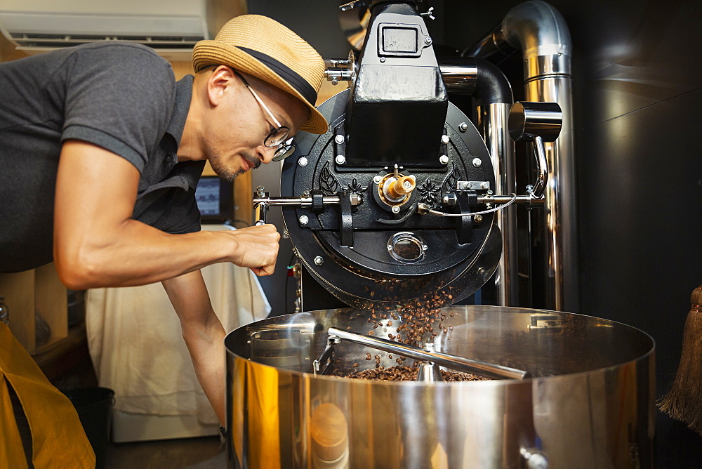 Japanese man wearing hat and glasses standing in an Eco Cafe, operating coffee roaster machine, Kyushu, Japan