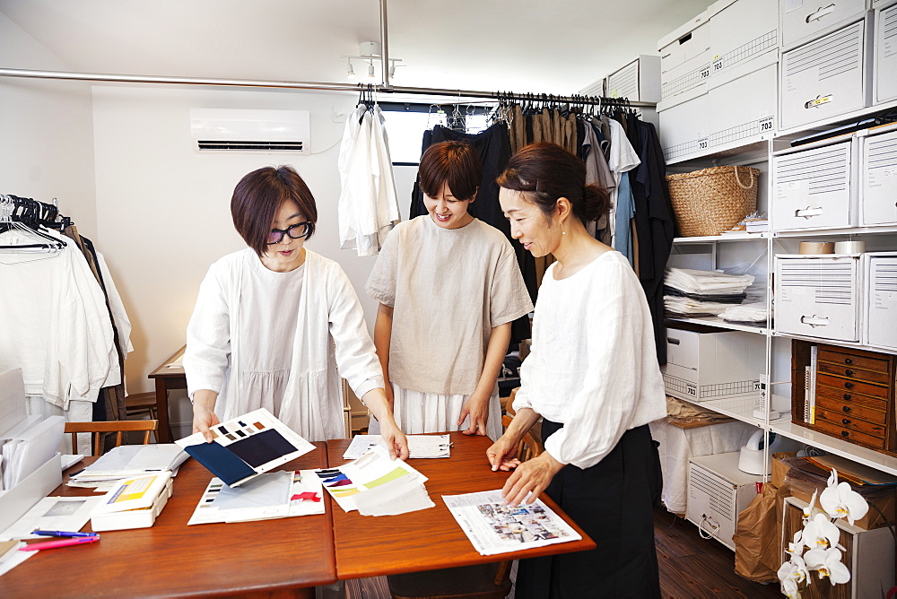 Three Japanese women standing at a table in a small fashion boutique, looking at fabric samples, Kyushu, Japan