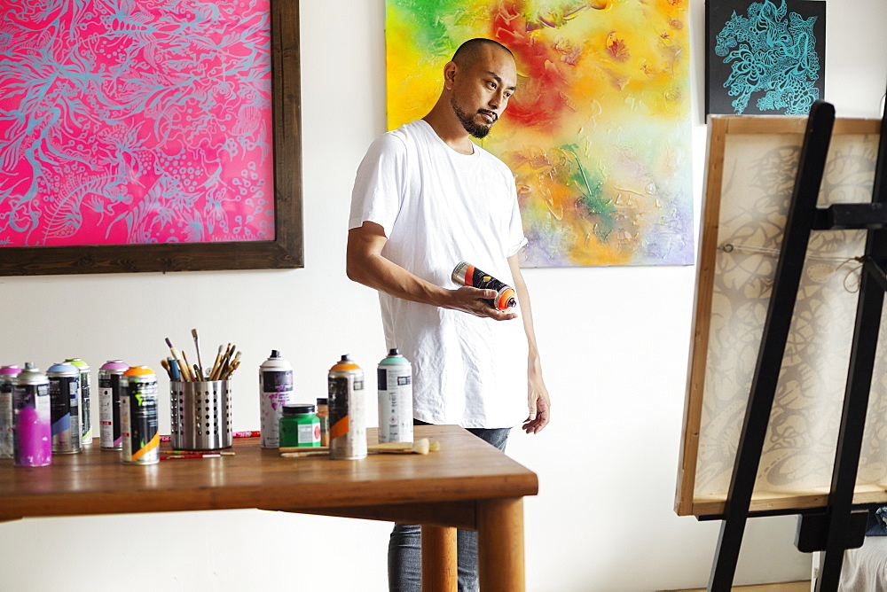 Japanese man standing in art gallery, holding can of spray paint, looking at artwork on easel, Kyushu, Japan