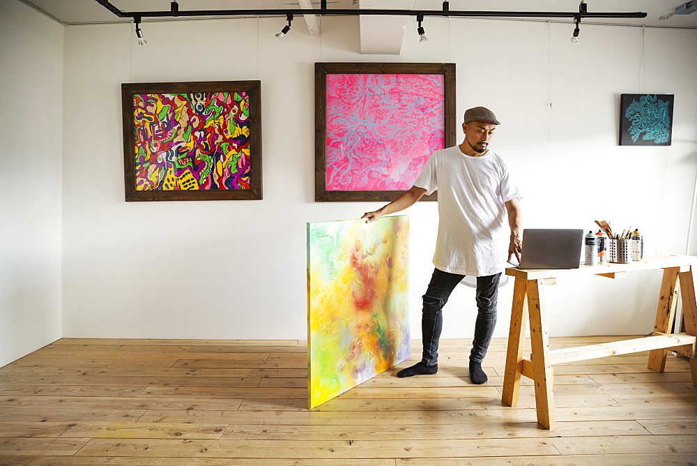 Japanese man standing in art gallery, holding abstract artwork, looking at laptop computer, Kyushu, Japan