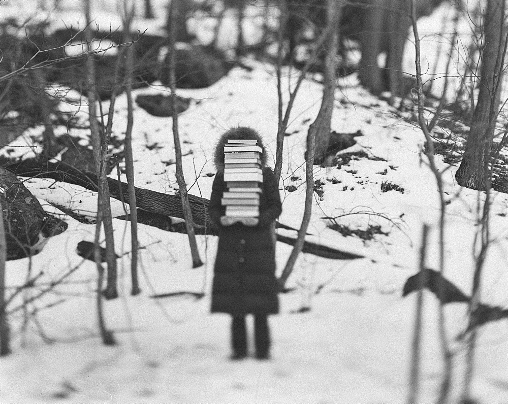 Child wearing a coat standing in a winter forest, holding a tall stack of books