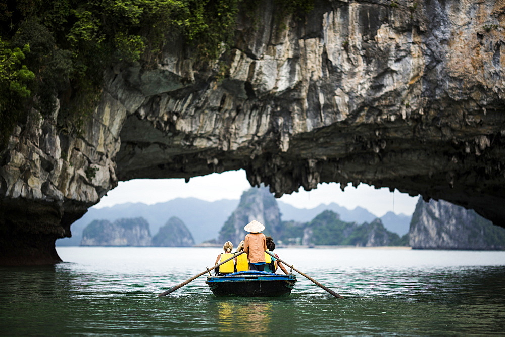 Rear view of man wearing straw hat transporting small group of people on a boat, rowing underneath natural rock arch, Vietnam