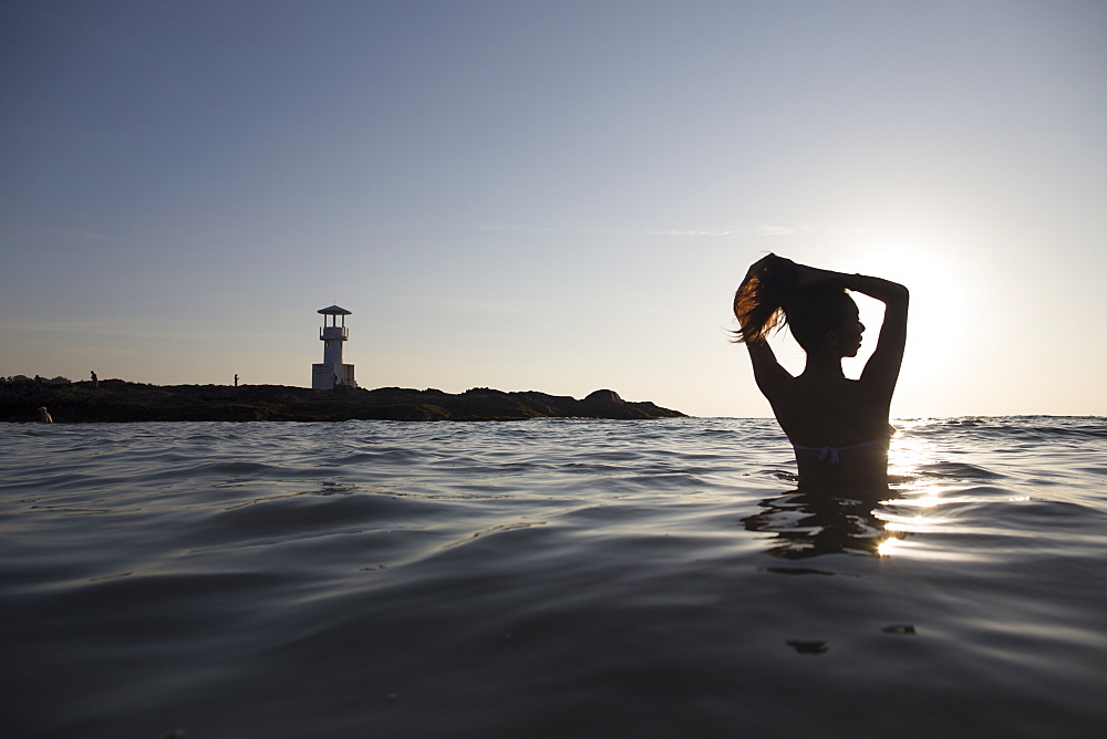 Rear view of woman bathing in the ocean at sunset, lighthouse in the distance