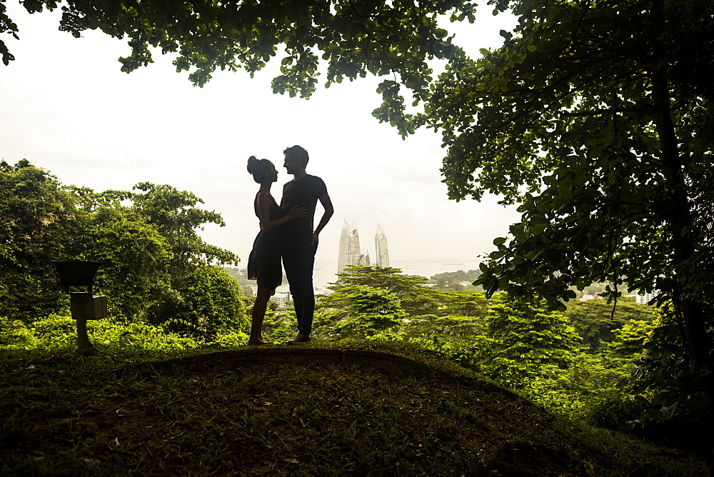 Silhouette of young couple standing underneath trees in a forest, skyscrapers in the distance, Singapore