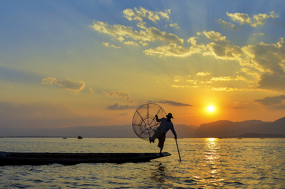 Traditional fisherman balancing on one leg on a boat, holding fishing basket, fishing on Lake Inle at sunset, Myanmar