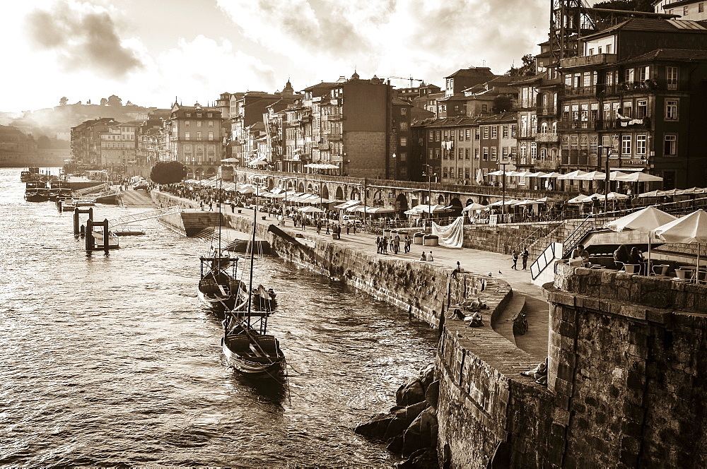 Port boats and freight barges moored beside a waterfront wall, and people on the promenade. Historic buildings, Porto, Portugal