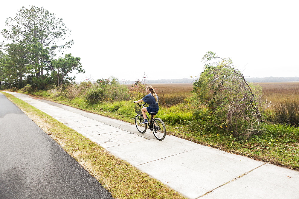 Teenage girl riding a bicycle along a path on open ground by water, St Simon's Island, Georgia, United States