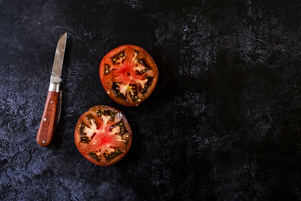 High angle close up of knife with wooden handle and fresh tomato cut in half and on black background, United Kingdom