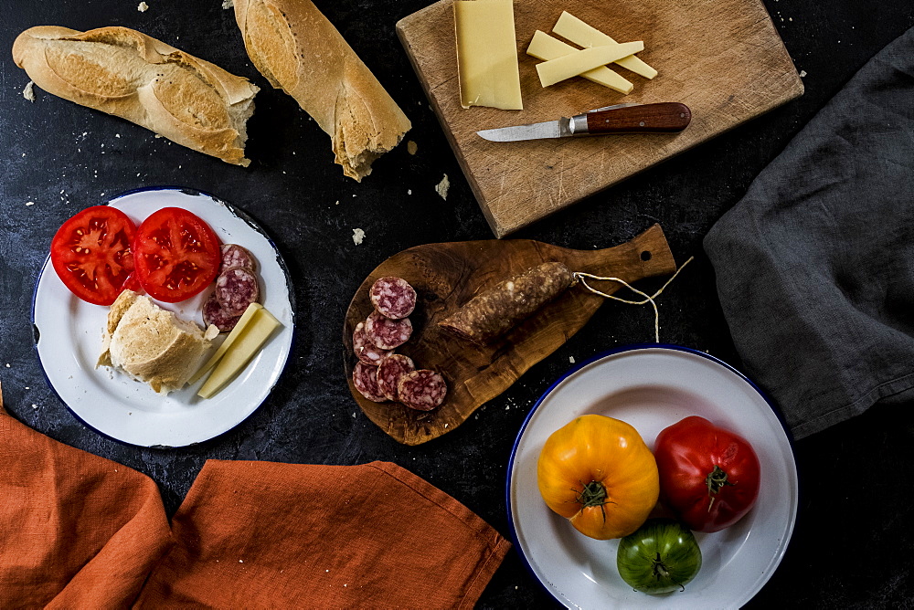 High angle close up of a selection of cheeses, tomatoes, salami and French baguette on white enamel plates on black background, United Kingdom