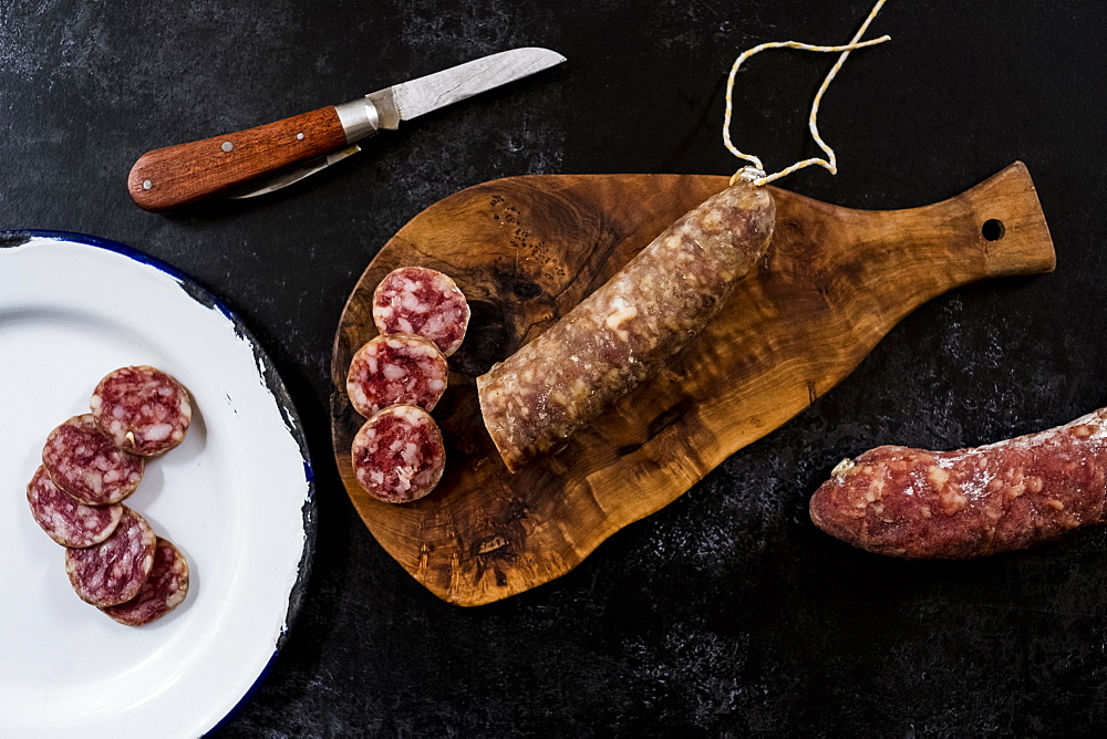 High angle close up of knife, sliced salami on wooden cutting board and white enamel plate on black background, United Kingdom