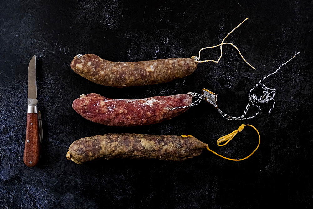 High angle close up of knife and selection of three salamis on black background, United Kingdom