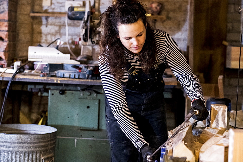 Woman with long brown hair wearing dungarees standing in wood workshop, using plane on piece of wood, Oxfordshire, United Kingdom