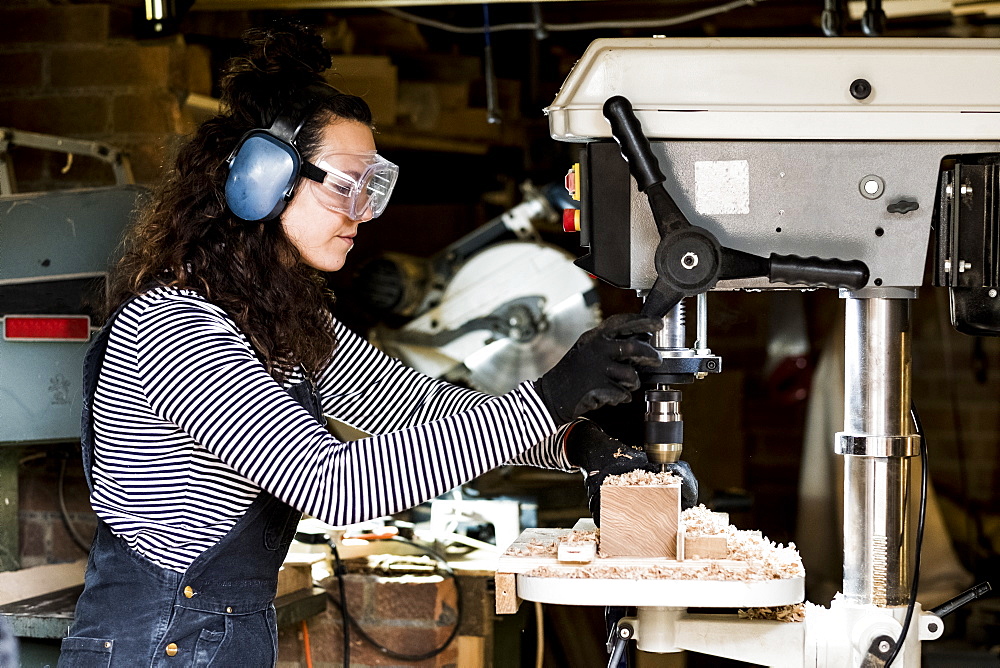 Woman with long brown hair wearing dungarees, safety glasses and ear protectors standing in wood workshop, using electric drill, Oxfordshire, United Kingdom